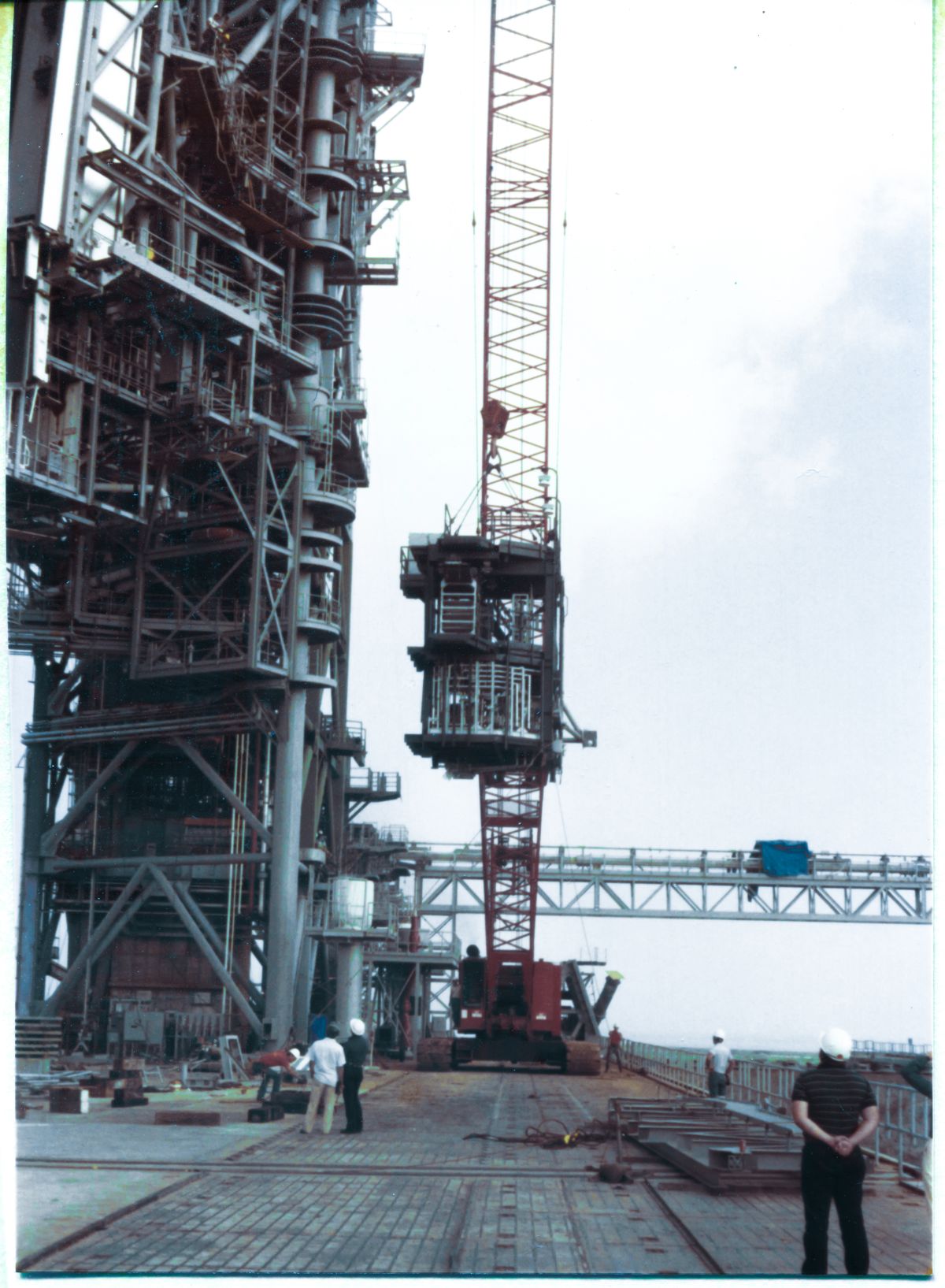Image 090. Having properly secured and verified that all is well with the suspended Orbiter Mid-Body Umbilical Unit, the crane operator has boomed left, just a little, and now has the OMBUU located out a little farther away from the face of the RSS, above the Crawlerway on the Pad Deck at Space Shuttle Launch Complex 39-B, Kennedy Space Center, Florida. He is now using only his load line to lift the hook which carries it aloft. A pair of Union Ironworkers continue to control the orientation of the OMBUU via the tag lines they will keep working for the duration of the lift, until the OMBUU reaches its destination. Up on the RSS, top left of frame, one of the connection plates which the bottom level of the OMBUU will bolt to, at RSS Column Line C-3 has come into view. To its right, the Access Catwalk at elevation 163'-9” has also come into view, and you can see ironworkers standing on it, ready to connect the OMBUU when it arrives. Beneath that catwalk, scaffolding is in place to give the pipefitters access to the run of plumbing which is supported beneath the catwalk, through which cryo and high-pressure gas will be fed into it from elevation 160'-0” on the FSS. Getting the OMBUU hung on the tower is only the beginning. A tremendous amount of work follows, getting it configured for its role supplying the Space Shuttle with consumables through the Mid-body Umbilical on the left side of the Orbiter's fuselage prior to lift-off. Photo by James MacLaren.